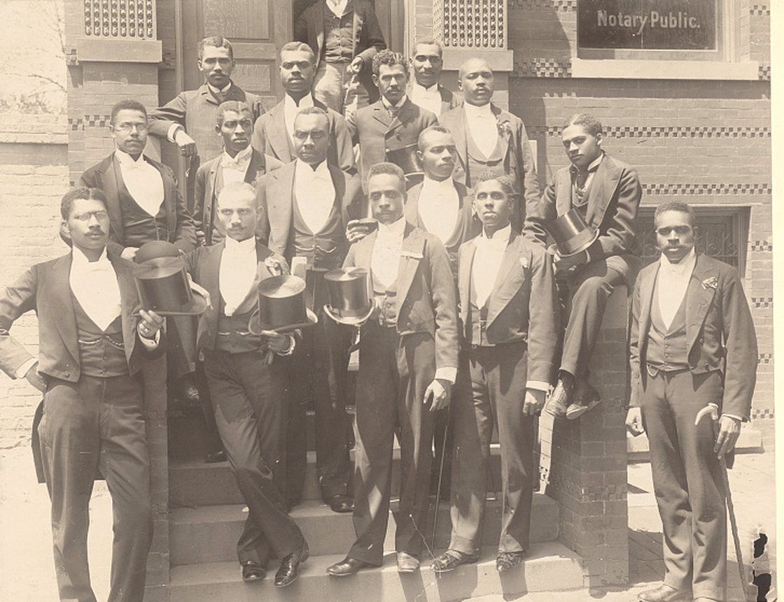 Sixteen Black law students in formal morning dress, several holding top hats and canes, on the steps of a building at Howard University about 1900. A Notary Public sign is visible in the window behind them.
