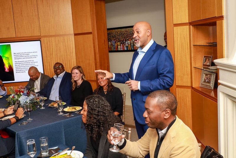 Roy L. Austin, Jr. addressing seated guests at a dinner, a slide describing the initiative on the screen behind him.