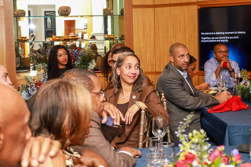 Guests seated around a dinner table listening, with a screen behind reading: We stand at a defining moment in history.